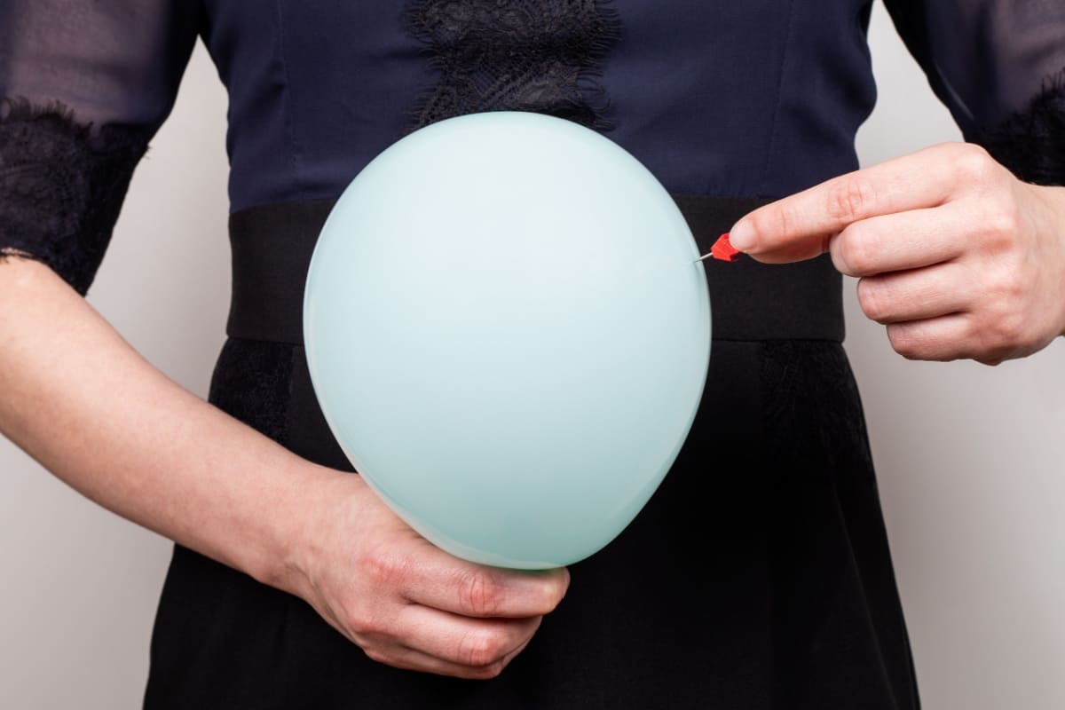 femeie sparge un balon tinut pe abdomen cu o pioneza 