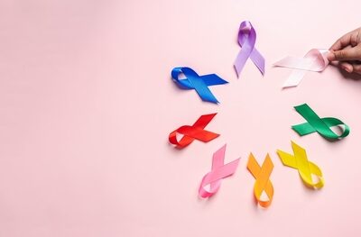 Cum te pregătești mental și fizic pentru o operație de cancer: sfaturi de la experți Mână ținând o panglică roz, așezată printre panglici colorate pe fundal roz, simbol al conștientizării cancerului și al altor cauze de sănătate.