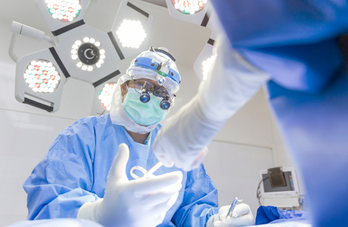 Chirurg bărbat în halat chirurgical albastru, aflat într-o sală modernă de cardiochirurgie, purtând ochelari loupă; medicul efectuează o intervenție pe cord, cu focalizare selectivă pe privirea chirurgului.