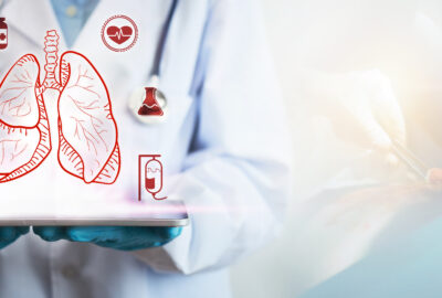 Cum să te pregătești pentru o intervenție chirurgicală toracică: ghid pentru pacienți și familii Concept de tratament al bolilor pulmonare, mâna medicului ține o tabletă care afișează o pictogramă a plămânilor, pe fundal fiind o imagine estompată dintr-o intervenție de chirurgie toracică.